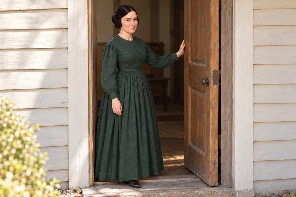 Emma Smith at thirty eight, wearing a dark green dress, stands in the open doorway of her home with one hand resting on the door.