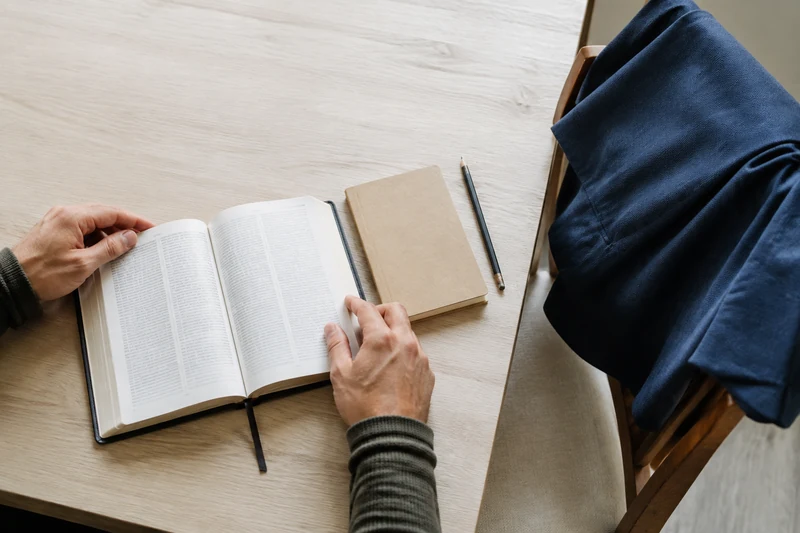 Hands holding an open scripture book beside a closed notebook and folded jacket