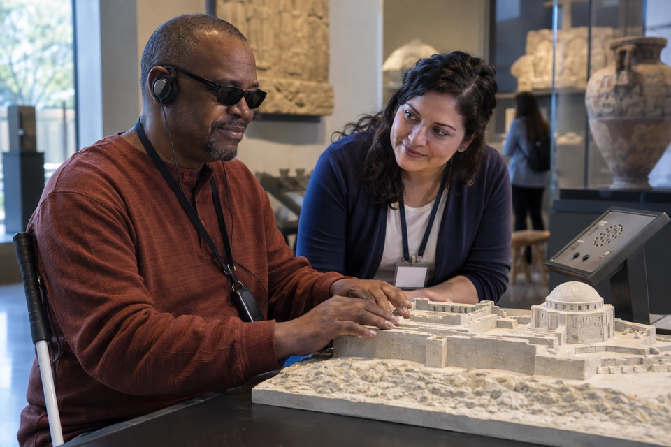 A man wearing headphones explores a raised architectural model with both hands while a woman watches attentively beside him.