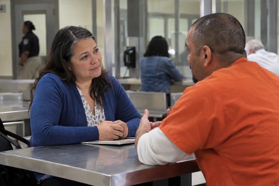 A woman listens across a metal table as a man in orange clothing speaks and gestures in an institutional visiting room.