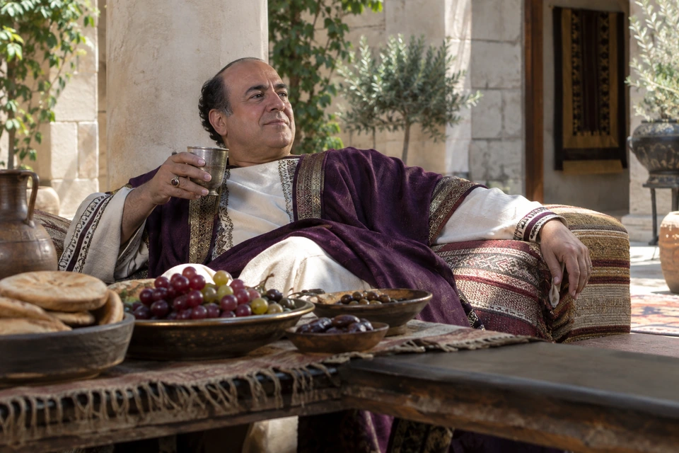 A man in purple and light linen reclines on cushions holding a metal cup beside a table laden with bread, fruit and olives.