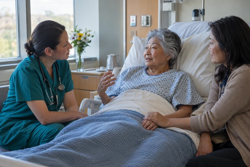 A nurse listens to an older patient while a family member sits beside the bed holding her hand.